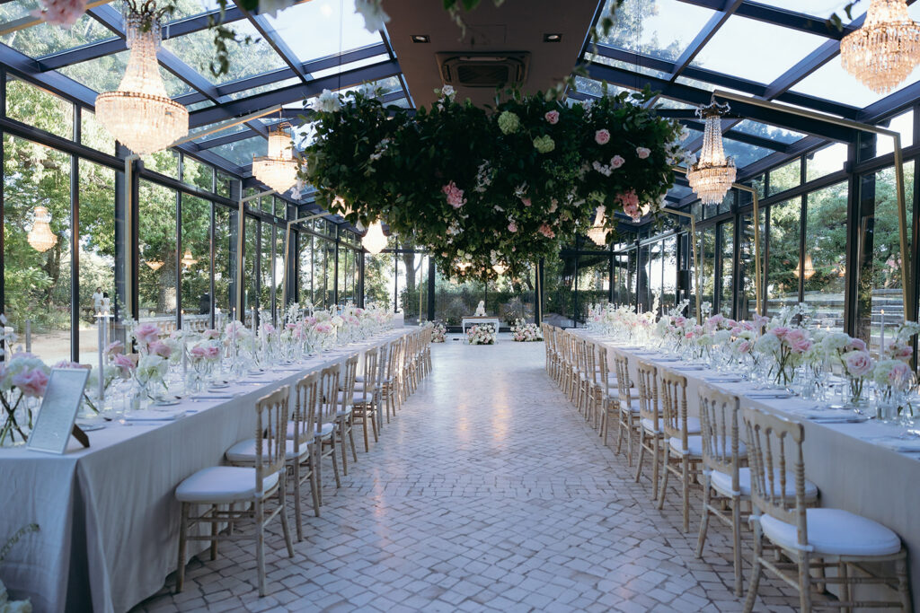 Two long wedding dining tables with a central aisle beneath hanging flowers and chandeliers in Vandelli’s glass-roofed reception space.