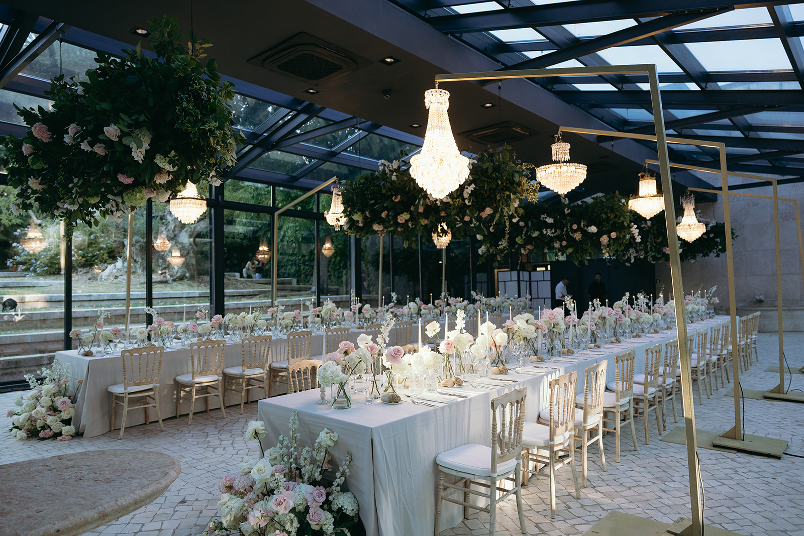 Wedding reception with long dining tables, hanging flowers and chandeliers beneath a glass roof.